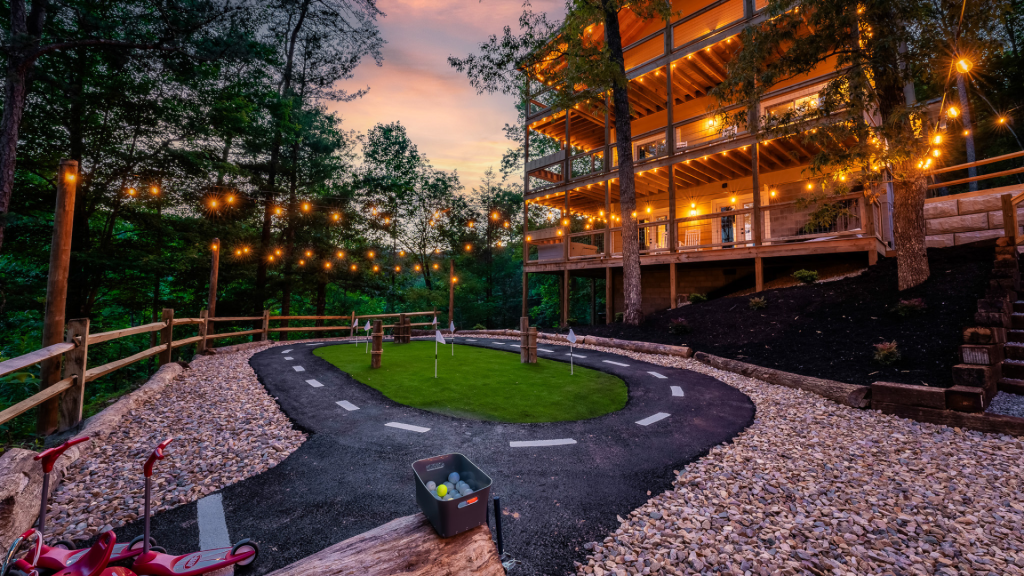 Mini golf course surrounded by string lights, next to a cozy multi-level cabin with multiple decks and private views.