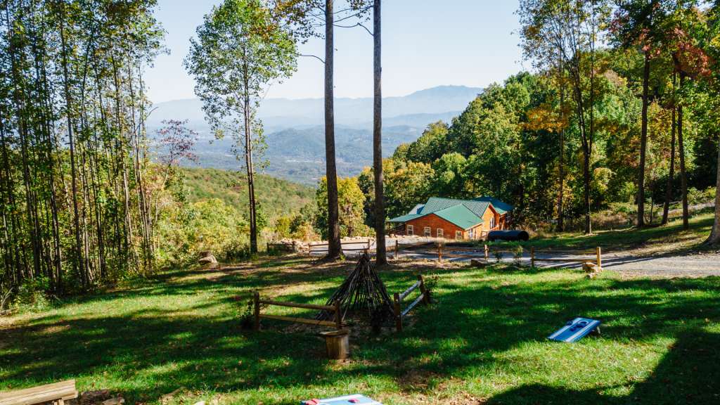 Mountain view with a cabin surrounded by trees and an outdoor play area featuring a cornhole game.