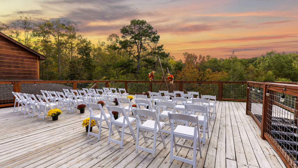 Wedding ceremony setup with white chairs and floral decorations on a wooden deck surrounded by trees.