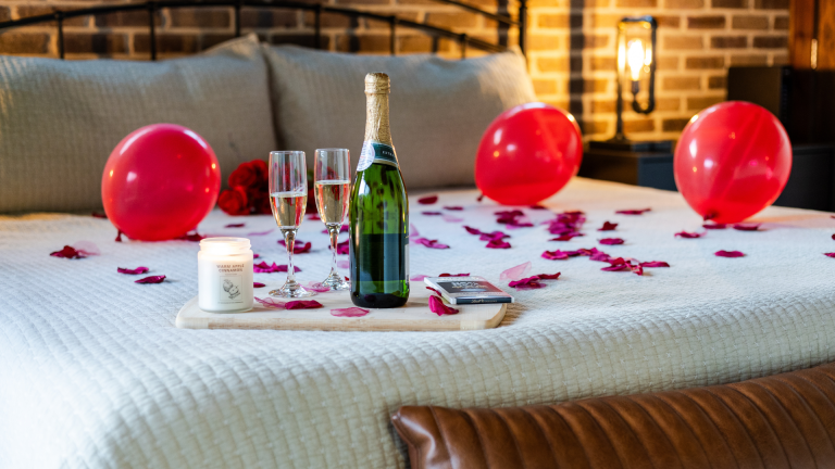 Romantic cabin bedroom setup with champagne, rose petals, red balloons, and candles for couples' getaways.