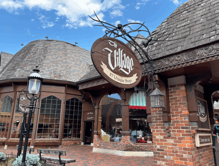 A rustic brick and wood building with a sign reading &ldquo;The Village: 27 Unique Shops.&rdquo; The curved shingle roofs and large windows evoke old-world charm, making the Village Shops Gatlinburg a must-see for memorable Gatlinburg shopping.