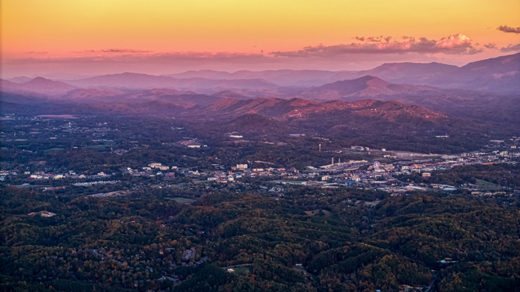 Aerial view of a town nestled in a lush green valley, surrounded by rolling hills and distant mountains under a pink and orange sunset sky.