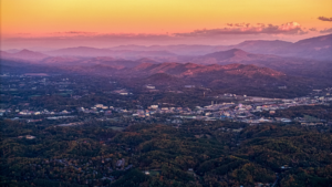 Aerial view of a town nestled in a lush green valley, surrounded by rolling hills and distant mountains under a pink and orange sunset sky.