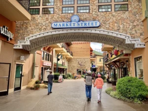 A stone archway labeled Market Street welcomes people into a lively outdoor shopping area designed with Elementor style, featuring boutique stores, plants, and string lights above the pathway. Three people are walking down the street.