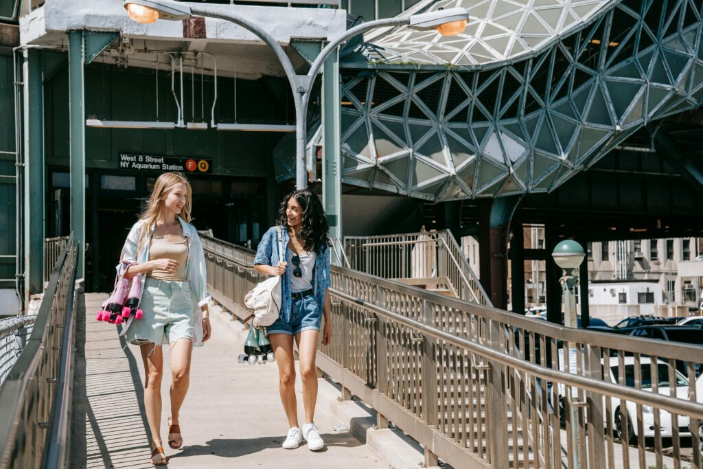 Two young women enjoy a sunny girls trip, walking and smiling together on a pedestrian ramp outside a subway station. One carries a yoga mat, and both have bags, embracing travel in an urban setting with modern architecture.
