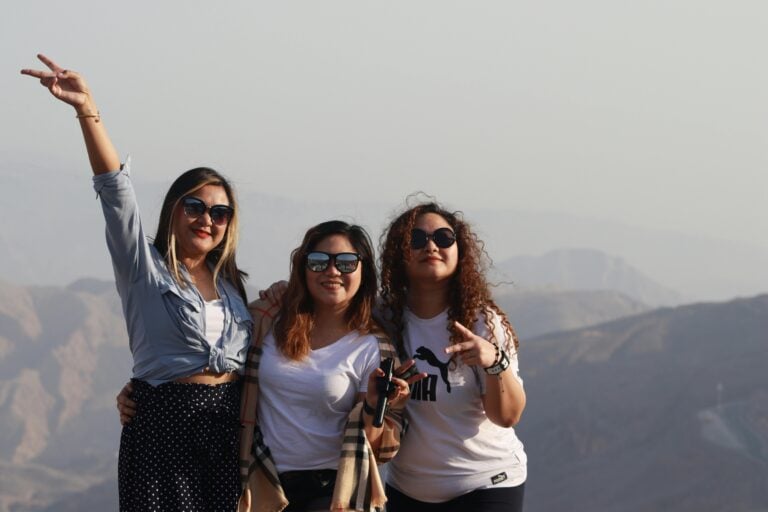 Three women stand outdoors in front of a mountainous landscape, smiling and posing for the camera. Two are making peace signs with their hands, all wearing sunglasses under a clear, bright sky—capturing an Elementor-worthy moment.