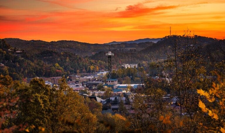 A scenic view of Gatlinburg nestled in the hills at sunset, with vibrant orange and yellow skies, scattered buildings, a tall observation tower, and autumn foliage—perfect for discovering new things to do in 2026.