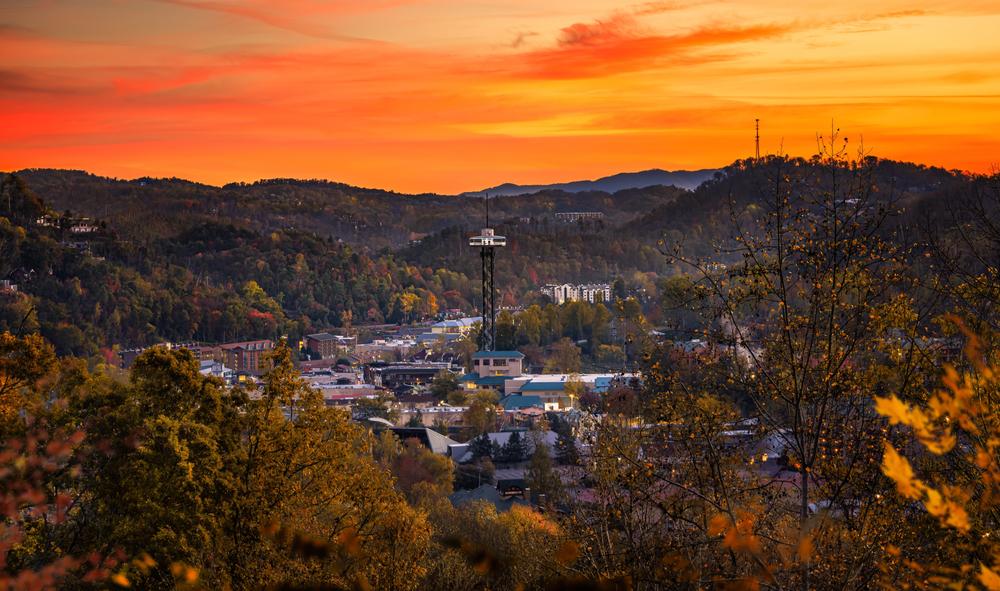 A scenic view of Gatlinburg nestled in the hills at sunset, with vibrant orange and yellow skies, scattered buildings, a tall observation tower, and autumn foliage—perfect for discovering new things to do in 2026.