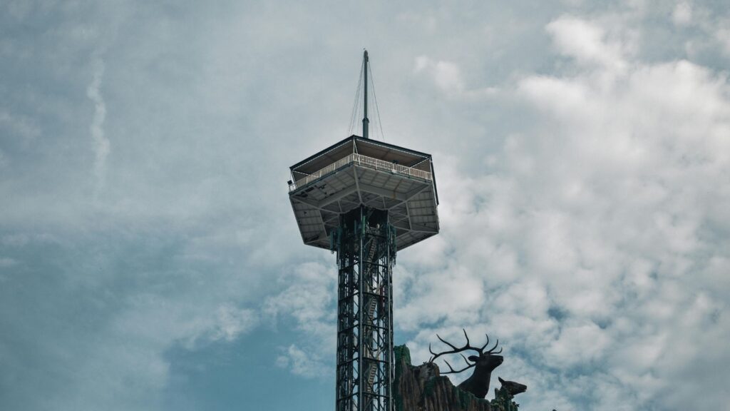 A tall observation tower with a pointed spire, the Gatlinburg Space Needle, rises against a cloudy sky; below, silhouettes of a large stag and small deer stand near the edge of a rocky structure—a classic scene among things to do in Gatlinburg.