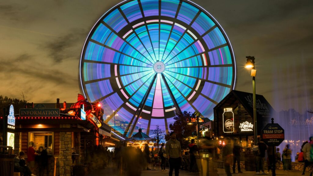 A large Ferris wheel with colorful, glowing lights spins above The Island in Pigeon Forge at dusk, where people stroll past illuminated signs and a nearby tram stop—one of the must-see Pigeon Forge attractions.