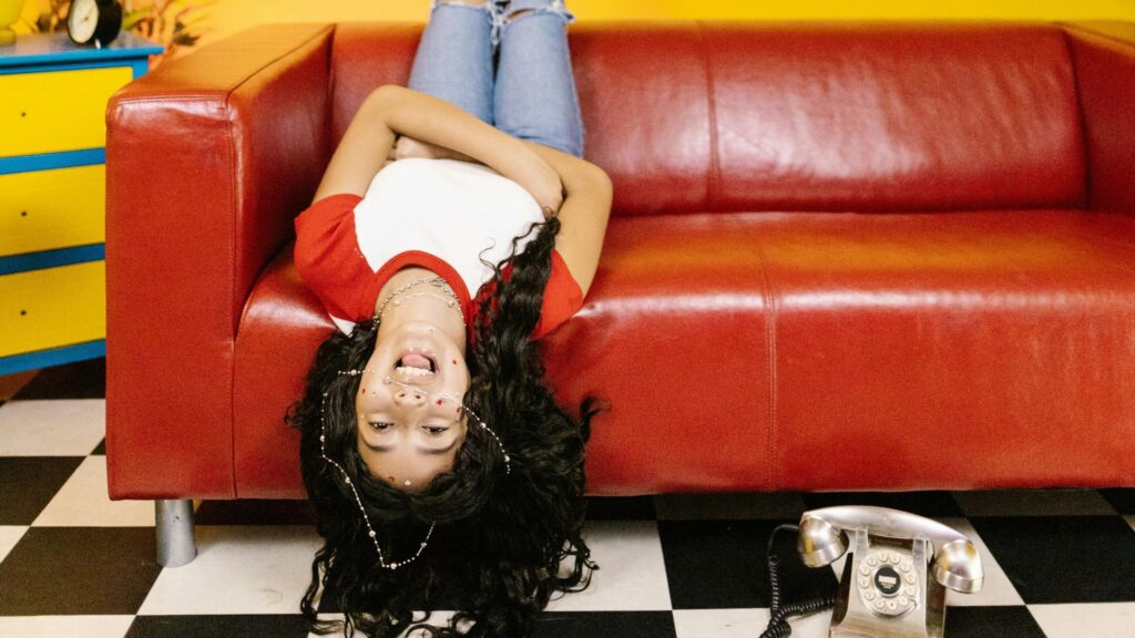 A person with long dark hair lies upside down on a red couch at WonderWorks Pigeon Forge, wearing a red and white shirt and blue jeans. The floor is black-and-white checkered, with an old-fashioned rotary phone nearby. Check hours and tickets before you visit!.