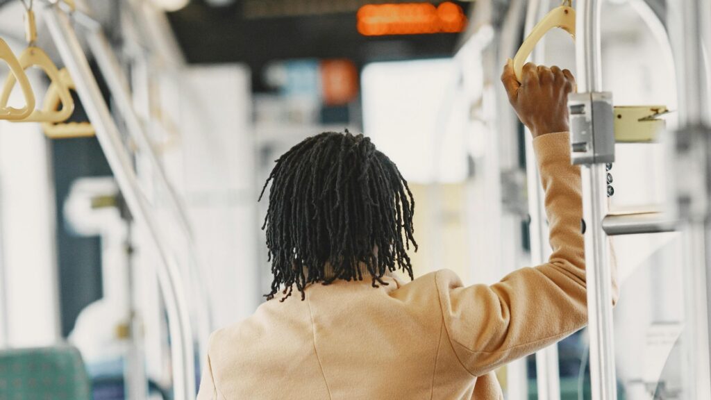 A person with short, twisted hair wearing a tan coat stands inside a Gatlinburg trolley, holding onto a metal handrail. The individual is facing away from the camera, experiencing convenient Gatlinburg transportation.