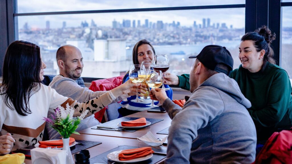 Five people sit around a table in a restaurant, smiling and clinking glasses for a toast, with a cityscape and water visible through large windows—perfect for large groups enjoying Gatlinburg dining.