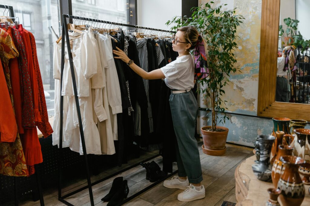 A woman wearing glasses, a white shirt, and jeans browses through clothes on a rack while shopping in Gatlinburg. The boutique features various garments, plants, and decorative items throughout the shop.
