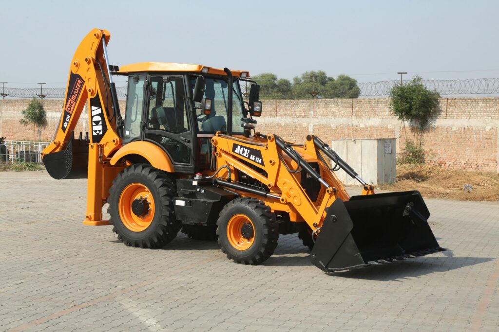 A yellow ACE backhoe loader is parked on a paved surface, featuring a front bucket and rear digging arm—just like the equipment you’ll see at Dig’n Zone Theme Park—with a brick wall and trees in the background.