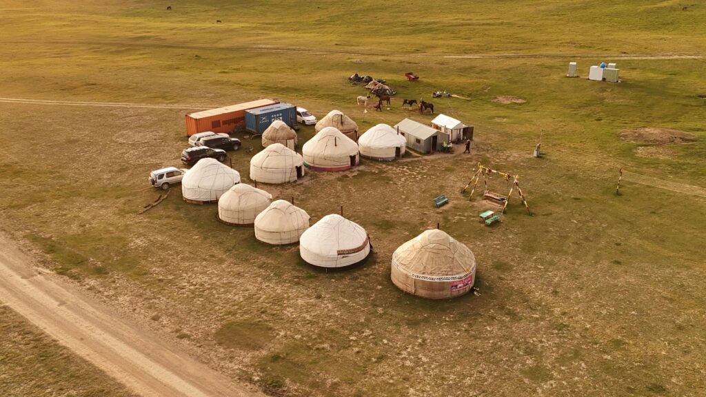 Aerial view of traditional yurts arranged in a row on grassland at campgrounds near Gatlinburg, with vehicles, horses, a shipping container, and portable toilets nearby. A dirt road runs along the edge of the camp.