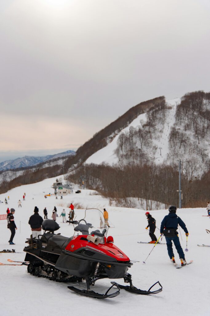 A red snowmobile is parked on the snowy ski slope at Ober Mountain in Gatlinburg, with a guide nearby as people ski and snowboard; snow-covered mountains and trees rise in the background under a cloudy sky.
