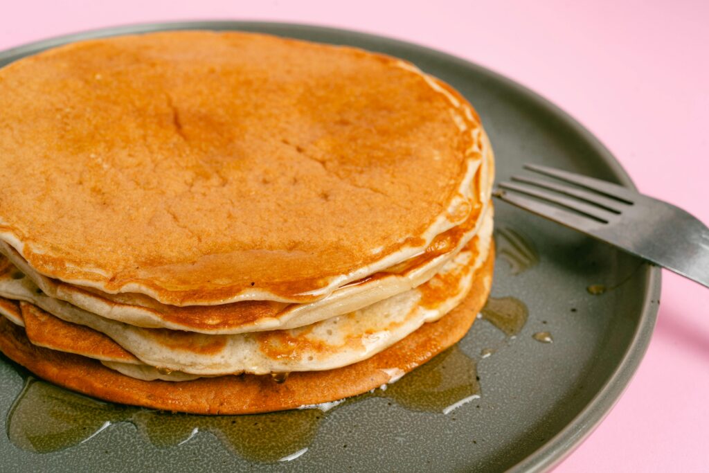 A stack of pancakes with syrup sits on a gray plate next to a fork, against a pink background, inspired by the menu at Crockett’s Breakfast Camp.