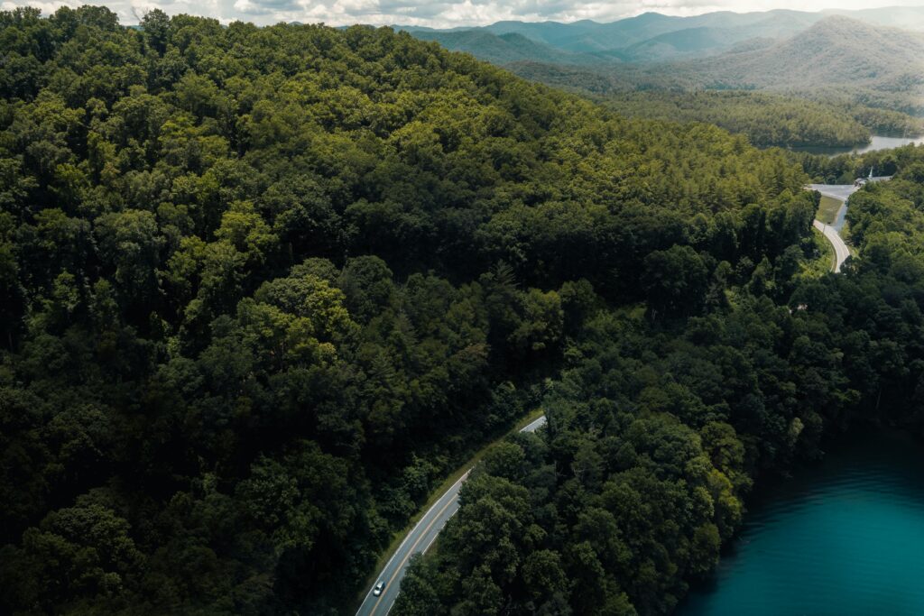 A winding road curves through dense, green forest beside a bright blue lake in Gatlinburg, offering scenic views of mountains rising in the background under a partly cloudy sky.