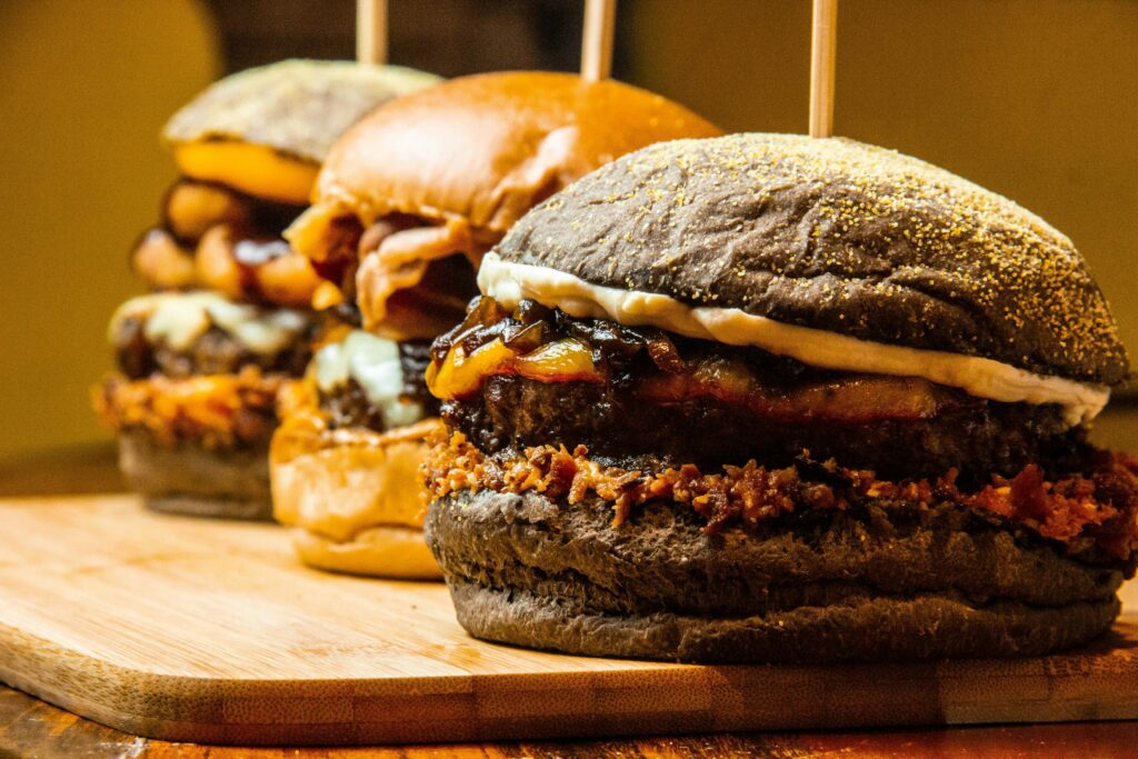 Three gourmet burgers with various toppings and sauces are arranged in a row on a wooden board&mdash;an enticing display reminiscent of top dining spots in the Smoky Mountains. The softly blurred background sets the perfect mood for dining in the Smokies.