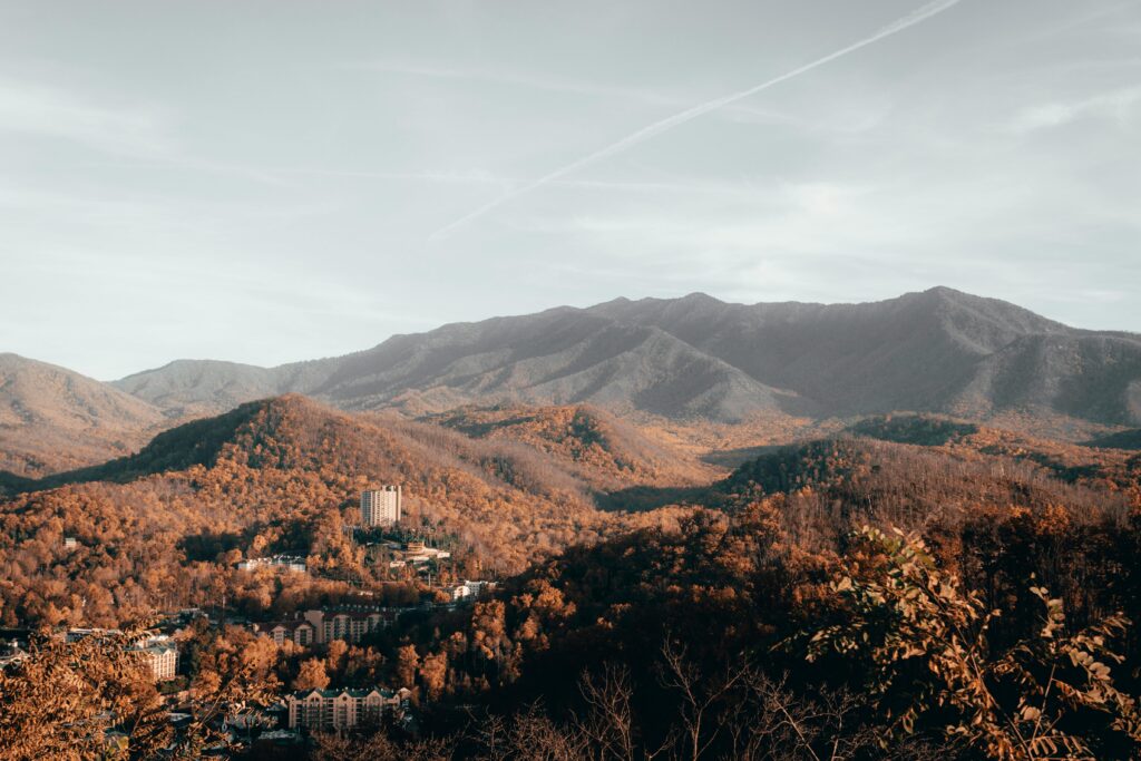 A scenic view of forested mountains with autumn foliage, scattered buildings, and a tall hotel structure nestled among the hills under a partly cloudy sky—one of Gatlinburg’s must see stops on any road trip.