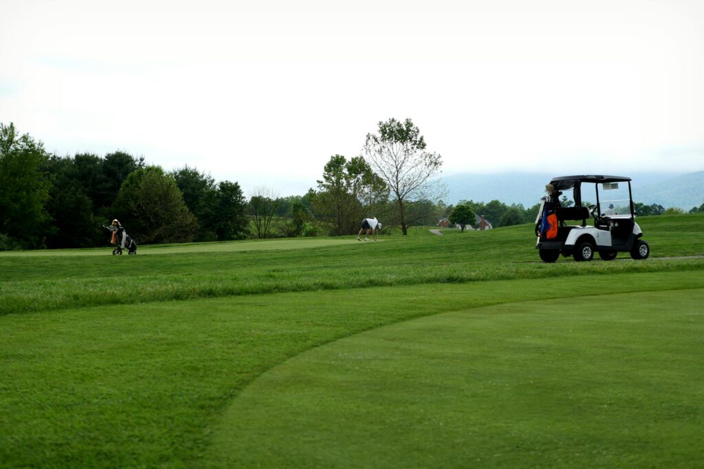 A golf course with several golfers playing in the distance. A white golf cart with clubs is parked on the grass near the right side of the image at Gatlinburg Golf Course, bordered by trees under a cloudy sky.