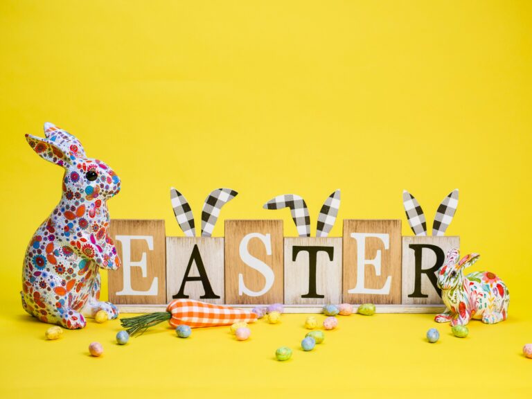 Colorful decorative rabbits and wooden blocks spelling EASTER with bunny ear decorations sit on a bright yellow background, surrounded by pastel candies and two fabric carrots—perfect inspiration for Smoky Mountains Easter activities.