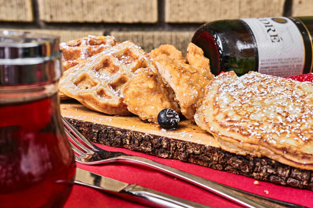 A close-up of waffles, pancakes, and fried chicken on a wooden board, perfect for those exploring the best places to eat Smokies. Powdered sugar and berries garnish the meal, with syrup and cutlery set on a red tablecloth.