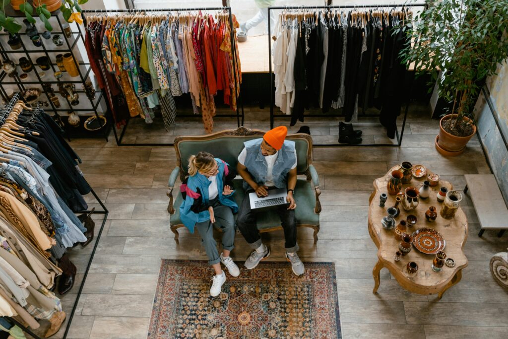 Two people sit on a couch in a vintage clothing store in Gatlinburg, one using a laptop. Racks of colorful clothes and a table with ceramics nearby create a cozy vibe—perfect for unique Gatlinburg shopping adventures. The scene is viewed from above.