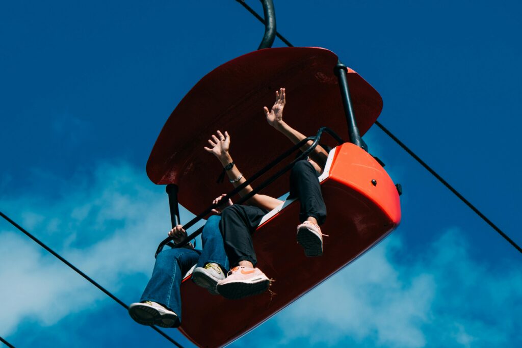 Two people sit in a bright red chairlift at Gatlinburg SkyPark against a blue sky, waving and dangling their legs as they enjoy the ride—a perfect moment when visiting Gatlinburg.