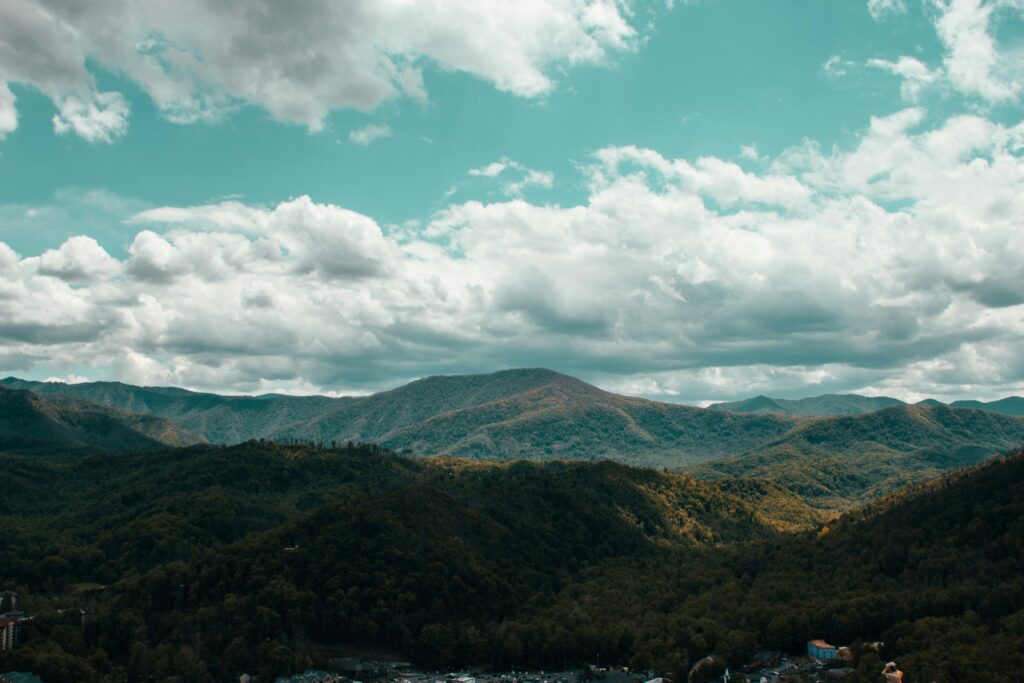 A scenic view of forested mountains near Gatlinburg, Tennessee, under a partly cloudy sky, with sunlight casting shadows across the rolling hills and valleys.