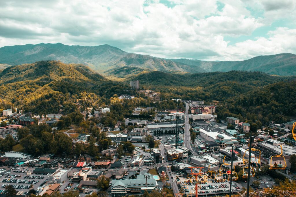Aerial view of Gatlinburg, Tennessee nestled in a lush green valley, surrounded by forested mountains under a partly cloudy sky. Buildings, roads, parked cars, and a glimpse of Anakeesta are visible in the foreground.