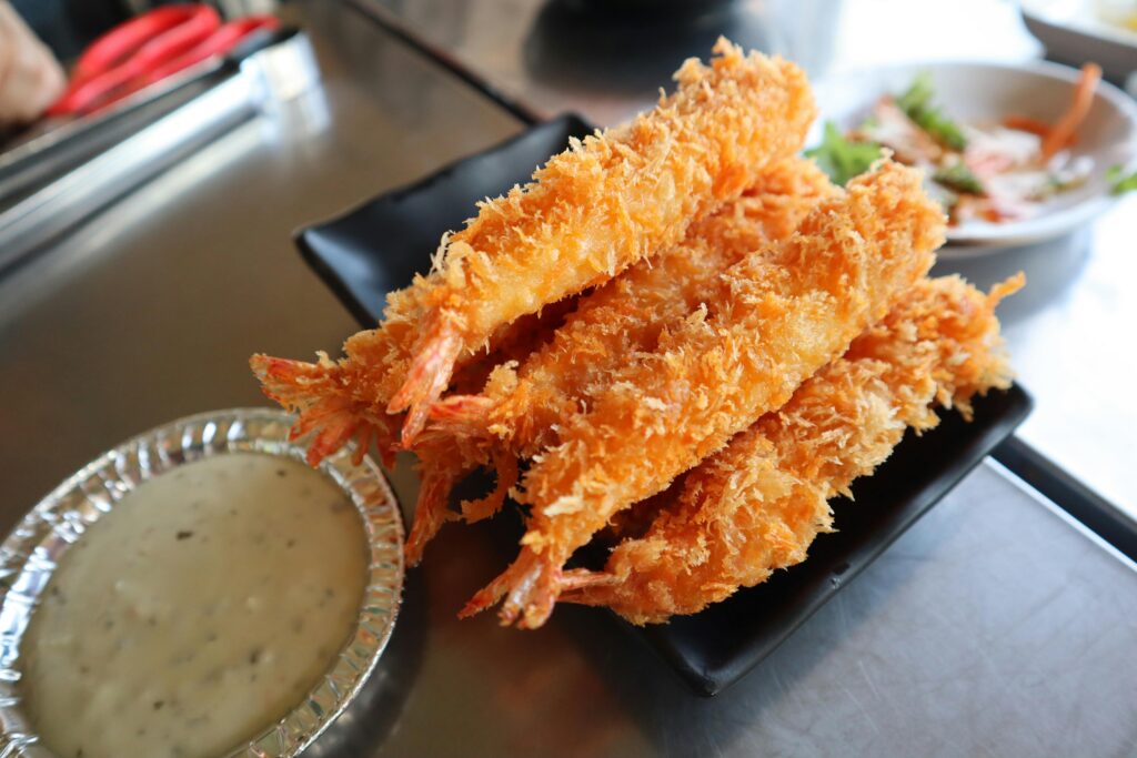 A black plate holds several pieces of golden, crispy fried shrimp from the Bubba Gump menu, with a foil dish of creamy dipping sauce nearby. In the background, there is a small plate of salad.