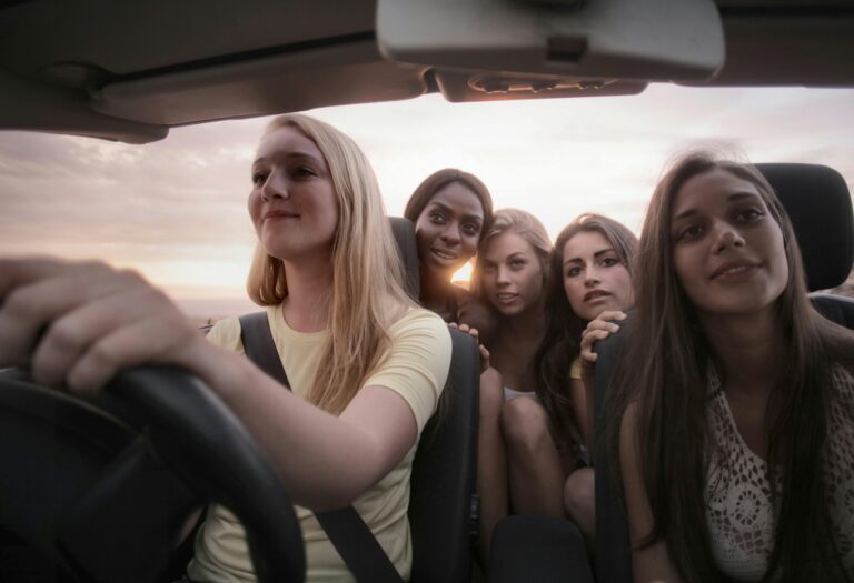 Five young women sit together in a car at sunset, with one driving and the others smiling and posing for a photo, capturing the cheerful spirit of a Girls’ Trip as they head toward one of the Best Places on their journey.