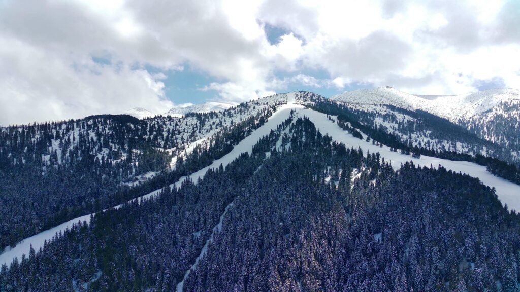A snow-covered mountain at Ober Mountain in Gatlinburg features pine trees and ski slopes under a partly cloudy sky. Sunlight filters through the clouds, illuminating the white peaks and dark green forest below—a perfect guide to winter beauty.