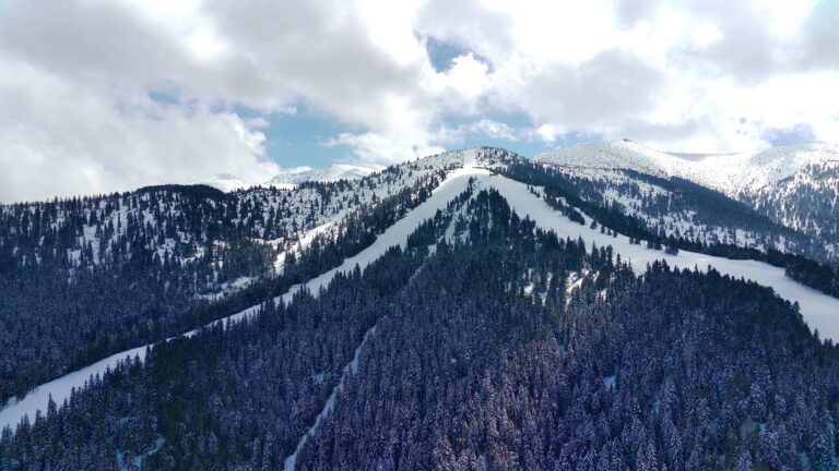 A snow-covered mountain at Ober Mountain in Gatlinburg features pine trees and ski slopes under a partly cloudy sky. Sunlight filters through the clouds, illuminating the white peaks and dark green forest below—a perfect guide to winter beauty.