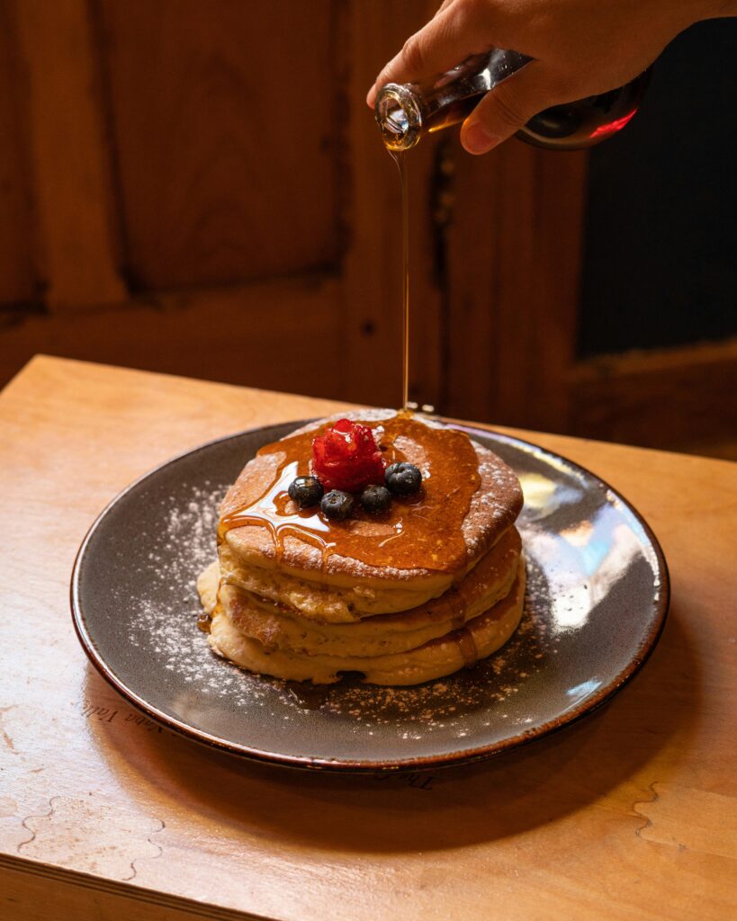 A hand pours syrup over a stack of pancakes topped with powdered sugar, blueberries, and a strawberry&mdash;an inviting breakfast served on a dark plate atop a wooden table, perfect for mornings in Gatlinburg.