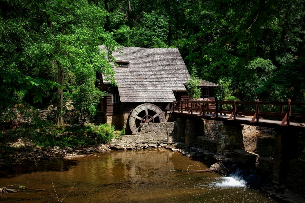 A rustic wooden watermill with a shingled roof sits by a stream, surrounded by dense green trees. Featured in many Old Mill Pigeon Forge guides, this scenic spot has a wooden bridge and sunlight filtering through the foliage.