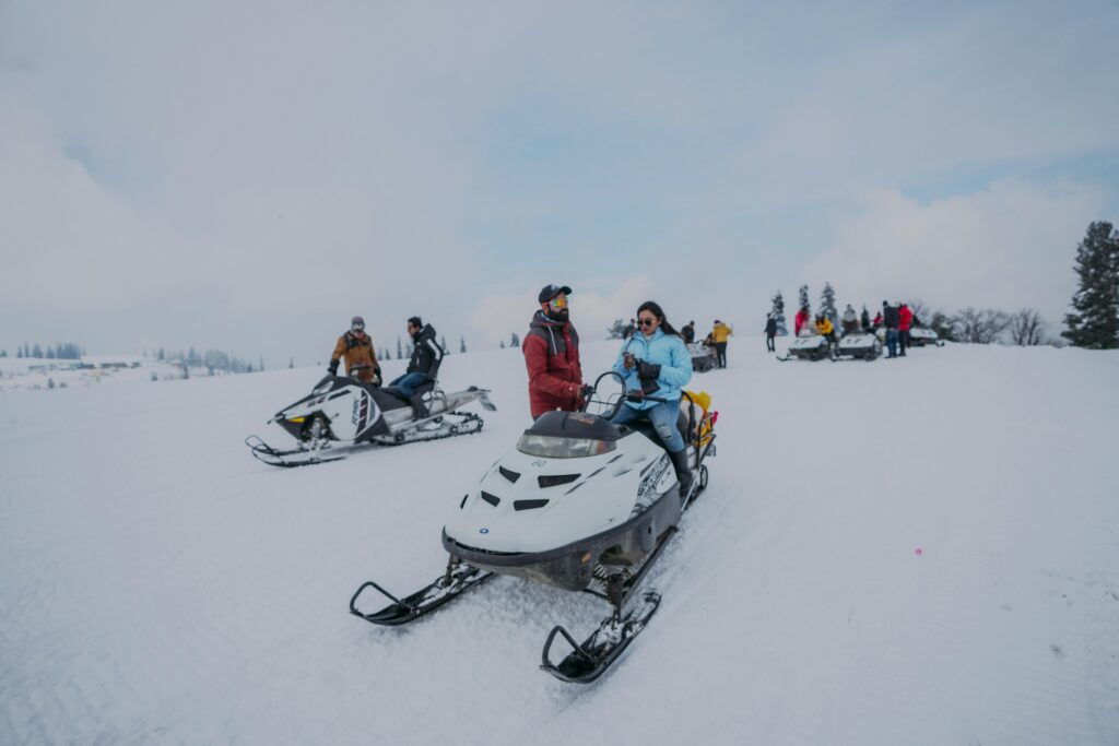 People enjoying winter activities on snowmobiles in a snowy landscape near Gatlinburg, TN. Dressed in winter clothing, some riders are moving while others are stationary among trees under a cloudy sky.