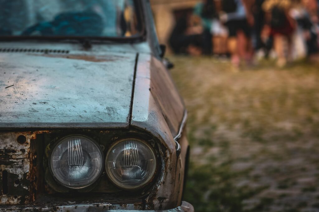 Close-up of the front left side of an old, rusty car with two round headlights, parked on a cobblestone road. The blurred background hints at people mingling, capturing the authentic vibe often found at Pigeon Forge car shows.