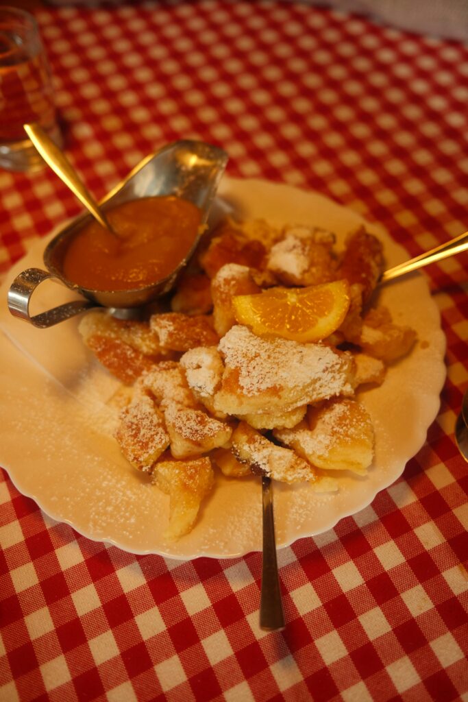 A plate of powdered sugar-dusted pancakes with an orange slice, served with applesauce on a red and white checkered tablecloth&mdash;classic comfort often found at top restaurants Smoky Mountains foodies love. Two forks rest on the plate.