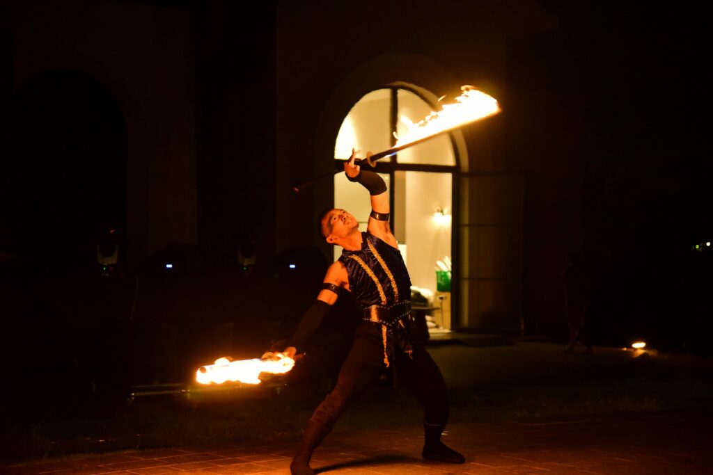 A fire performer in dark clothing dramatically poses at night, holding two flaming batons—one above his head and one extended outward—against the backdrop of an arched doorway, reminiscent of the thrilling Dinner Shows in Pigeon Forge.
