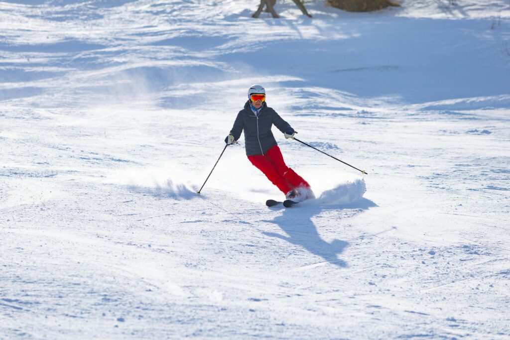 A person wearing a helmet, goggles, a dark jacket, and bright red pants is skiing downhill on a snowy slope at Ober Mountain in Gatlinburg, creating a spray of snow as they make a turn under bright sunlight.