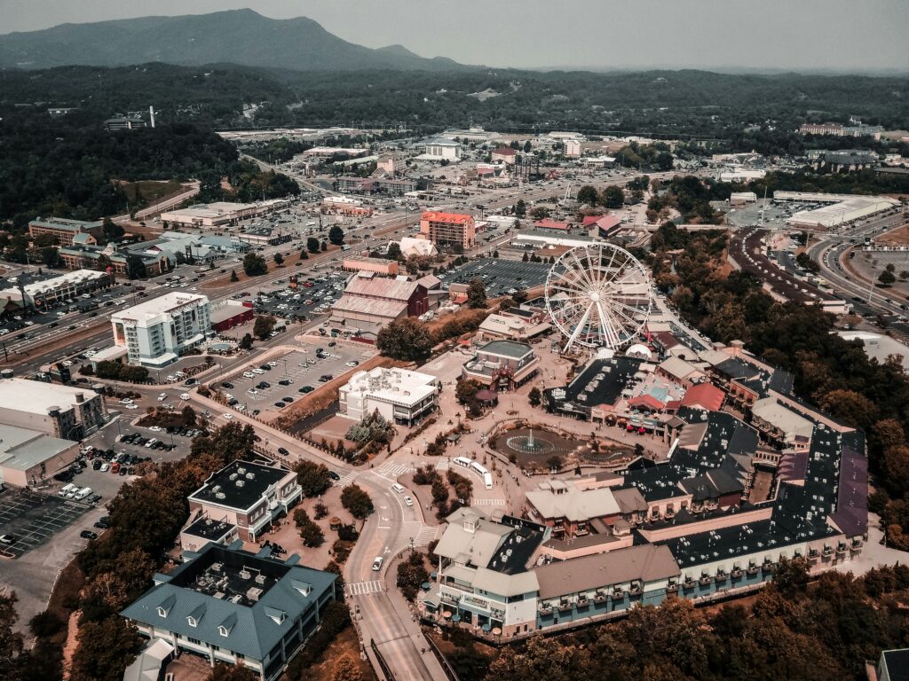 Aerial view of Pigeon Forge, showcasing its iconic Ferris wheel at the center, surrounded by city attractions, buildings, parking lots, roads, and distant green hills under a hazy sky.