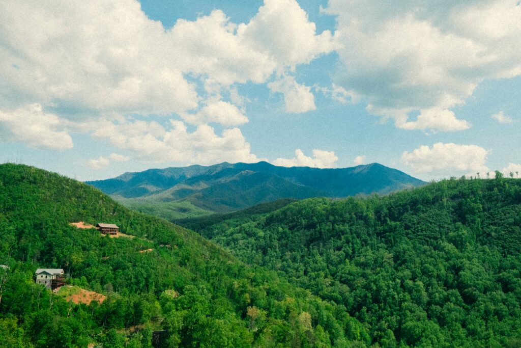 A scenic view of lush green forested hills near Gatlinburg, Tennessee, with a few houses scattered, set against a distant range of blue mountains under a partly cloudy sky.