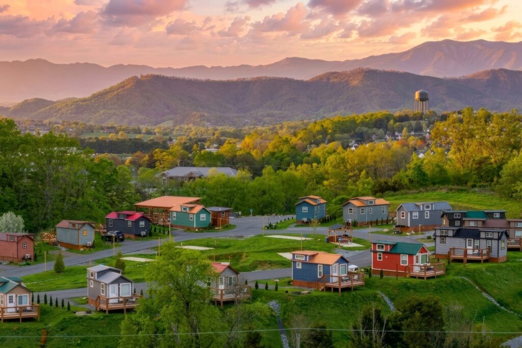A colorful tiny house village sits among green trees and rolling hills at sunset, with Smoky Mountain peaks and a rustic water tower in the background, evoking the charm of cozy cabin communities.
