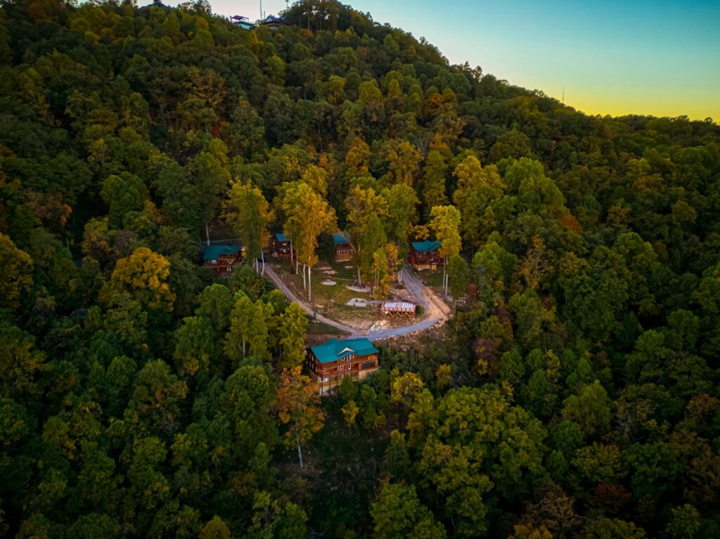 Aerial view of cabins nestled among dense, green forest on a hillside at sunset, with clearings and a winding dirt path connecting the buildings.