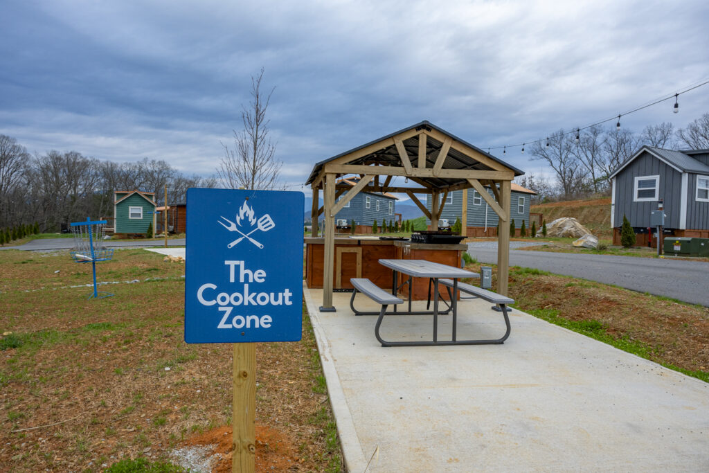 A sign reading The Cookout Zone stands by a covered picnic table and grill area on a concrete pad, surrounded by grassy open space and small houses under a cloudy sky—evoking the relaxed charm of Smoky Mountain cabin communities.
