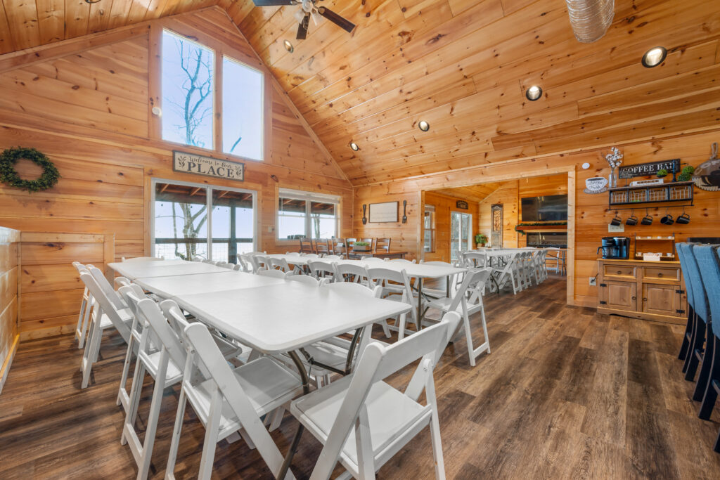 Bright, spacious, and ready for gatherings or events, this large wooden room with a vaulted ceiling features several long white folding tables and chairs, large windows, a coffee station, and rustic décor—embodying the inviting charm found in Smoky Mountain Cabins.