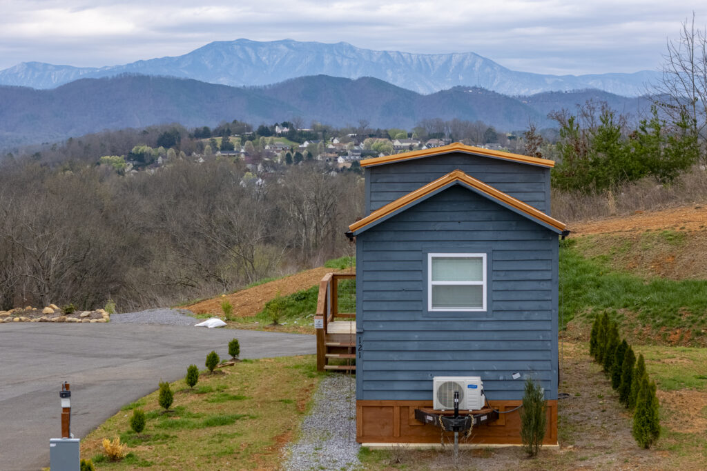 A small blue cabin sits on a grassy lot with the Smoky Mountain range and a town visible in the distance under a cloudy sky. Shrubs are planted along the path beside the house, creating a cozy atmosphere perfect for those who cherish close-knit communities.